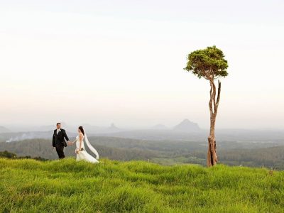 A bride and groom standing on a grassy hill with a tree in the background on the Sunshine Coast.