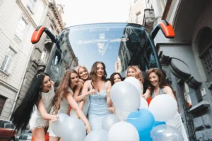Group of joyful women in elegant dresses celebrating with wedding transport balloons near a bus on a city street.