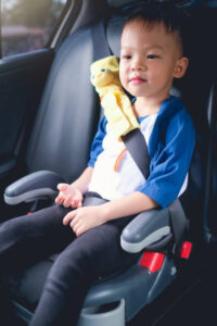 A young boy with a toy duck on his arm is seated in a child safety car seat, looking to the side with a slight smile, as he waits for airport transfers. The car interior is visible