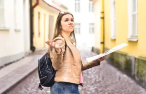 A confused young woman holding a map and looking up, standing on a street lined with colorful buildings. She wears a tan jacket, denim jeans, and carries a backpack, seemingly figuring out airport transfers.