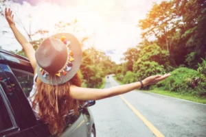 A woman wearing a straw hat decorated with flowers sticks her arms out of a car window, expressing joy on a scenic road surrounded by lush greenery during her airport transfer.