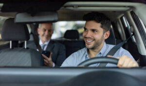 A young man smiling while driving a car, with an older man in business attire using a smartphone in the back seat. They appear to be in a comfortable, well-lit vehicle, possibly en route