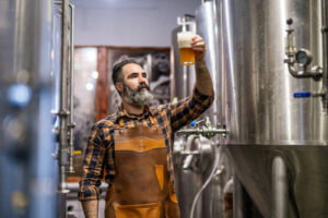 A bearded man in a plaid shirt and apron examines a glass of beer in a brewery with stainless steel tanks in the background during a Food & Wine Tour.