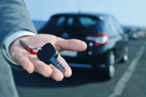 A person's hand in a suit offering a car key with a red key tag, displaying transport options in Noosa, with a blurred black car in the background on a sunny day.