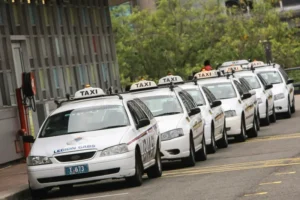 A line of white legion cabs taxis parked along a city street in Noosa, each with a "taxi" sign on top, waiting by the curb with greenery in the background.
