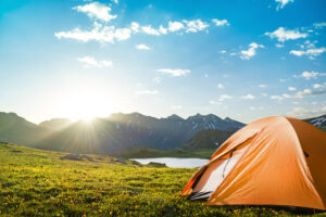 An orange tent pitched on a grassy meadow with wildflowers, overlooking a serene lake surrounded by rugged mountains. The scene is illuminated by the warm glow of a setting sun while holidaying on the