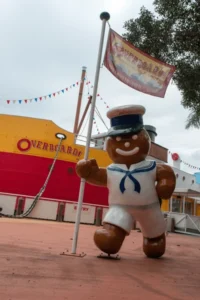 A large statue of a gingerbread man in a sailor outfit, holding a flagpole with a sign that reads "Why Visit the Sunshine Coast?" stands in front of a yellow and red ferry boat named