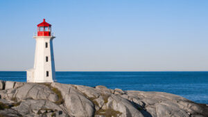 A white lighthouse with a red top stands on rugged, gray rocks against a backdrop of a calm blue sea and clear sky, making it one of the top destinations to visit on the Sunshine Coast.